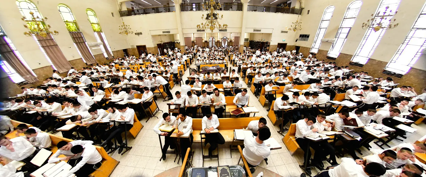 A bright beis medrash filled with learners beneath stained-glass windows